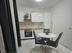 A compact kitchen with white cabinets, a built-in oven, and a round dining table with chairs. - Mary Apartments & Pool (Trogir)