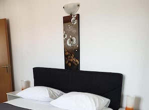 A bed with white linens, a dark headboard, and a wall-mounted light fixture with a floral design. - Mary Apartments & Pool (Trogir)