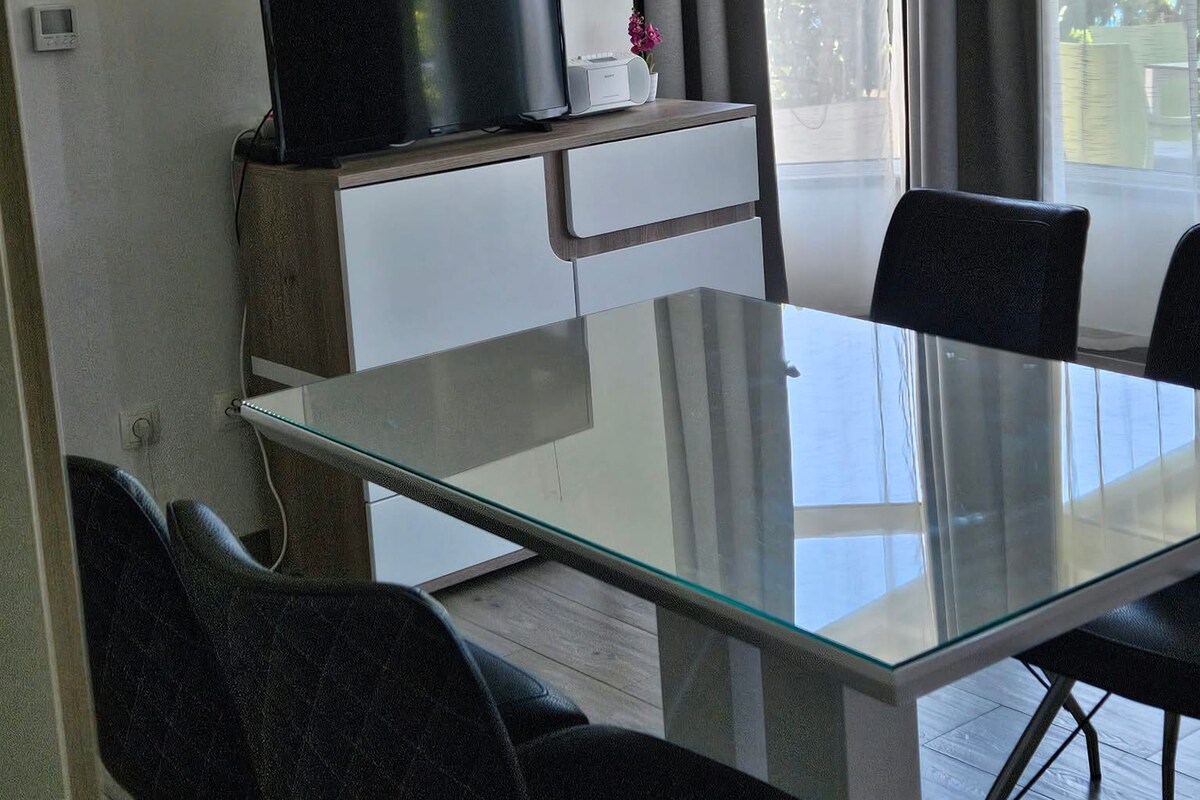 A modern dining area with a glass table and black chairs, a small cabinet with a coffee machine, and a view of the outdoors through a window.