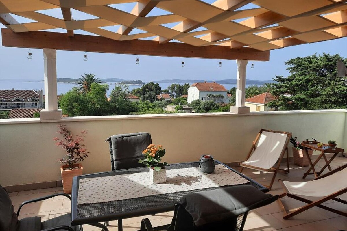 A covered outdoor seating area with a table and chairs, overlooking a coastal landscape.