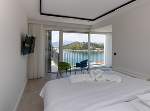 A hotel room with a bed, a TV, a balcony with chairs, and a scenic view of a lake and mountains.