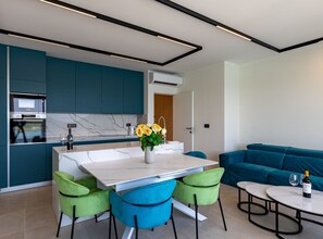 A modern kitchen with built-in appliances, a dining area with a white table and green chairs, and a teal-colored sofa in the living area.