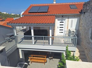 A two-story building with a red-tiled roof, a balcony with a metal railing, and a small garden area with a bench and potted plants. - Villa Lili (Murter-Kornati)