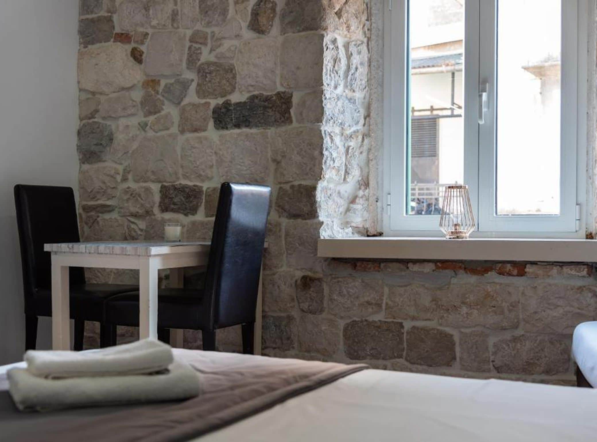 A room with a stone wall, a window with a view, a bed with a blanket, a chair, and a table.