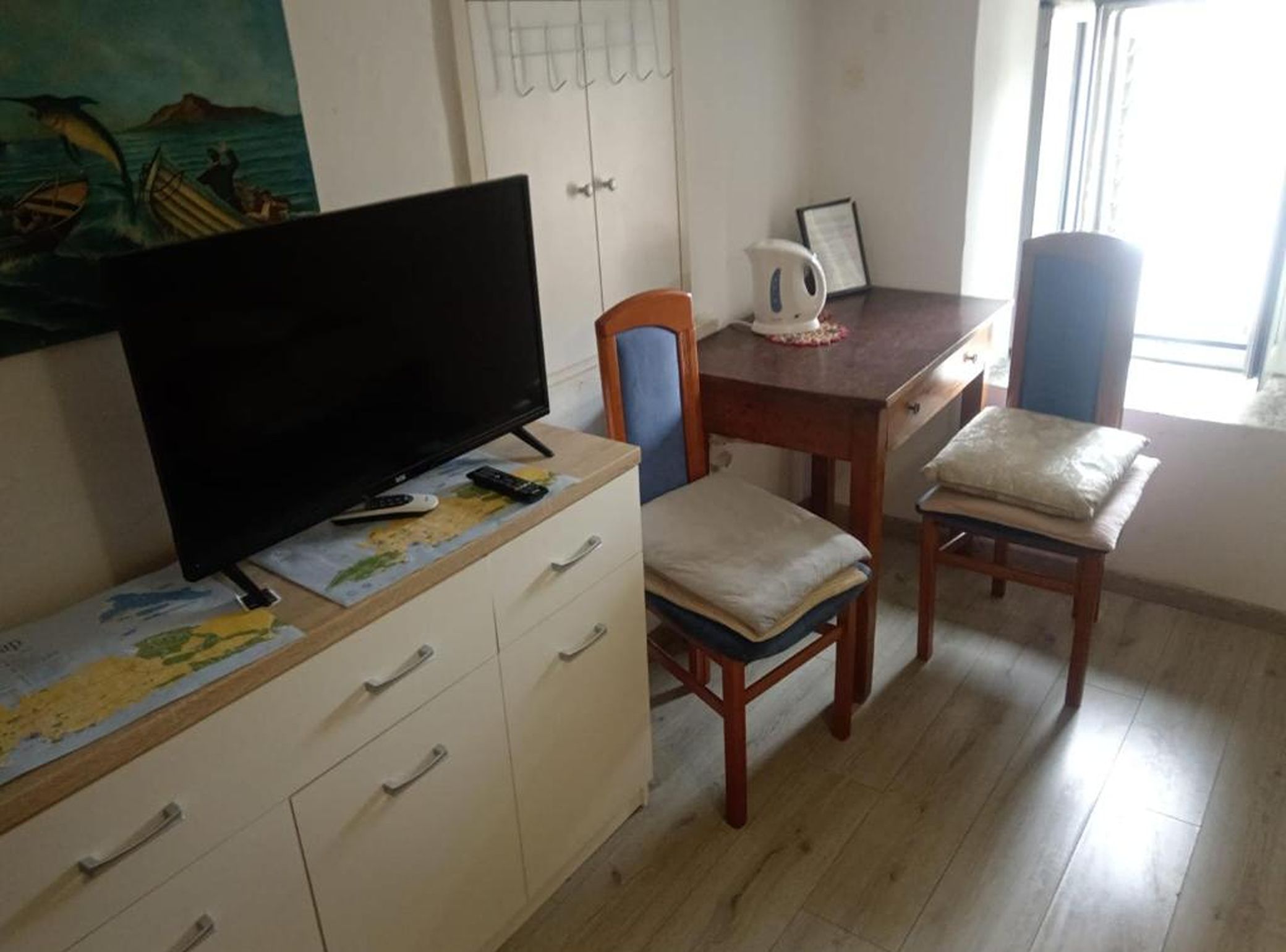 A room with a flat-screen TV on a dresser, a map, and a coffee table with a kettle. There are two wooden chairs and a table.