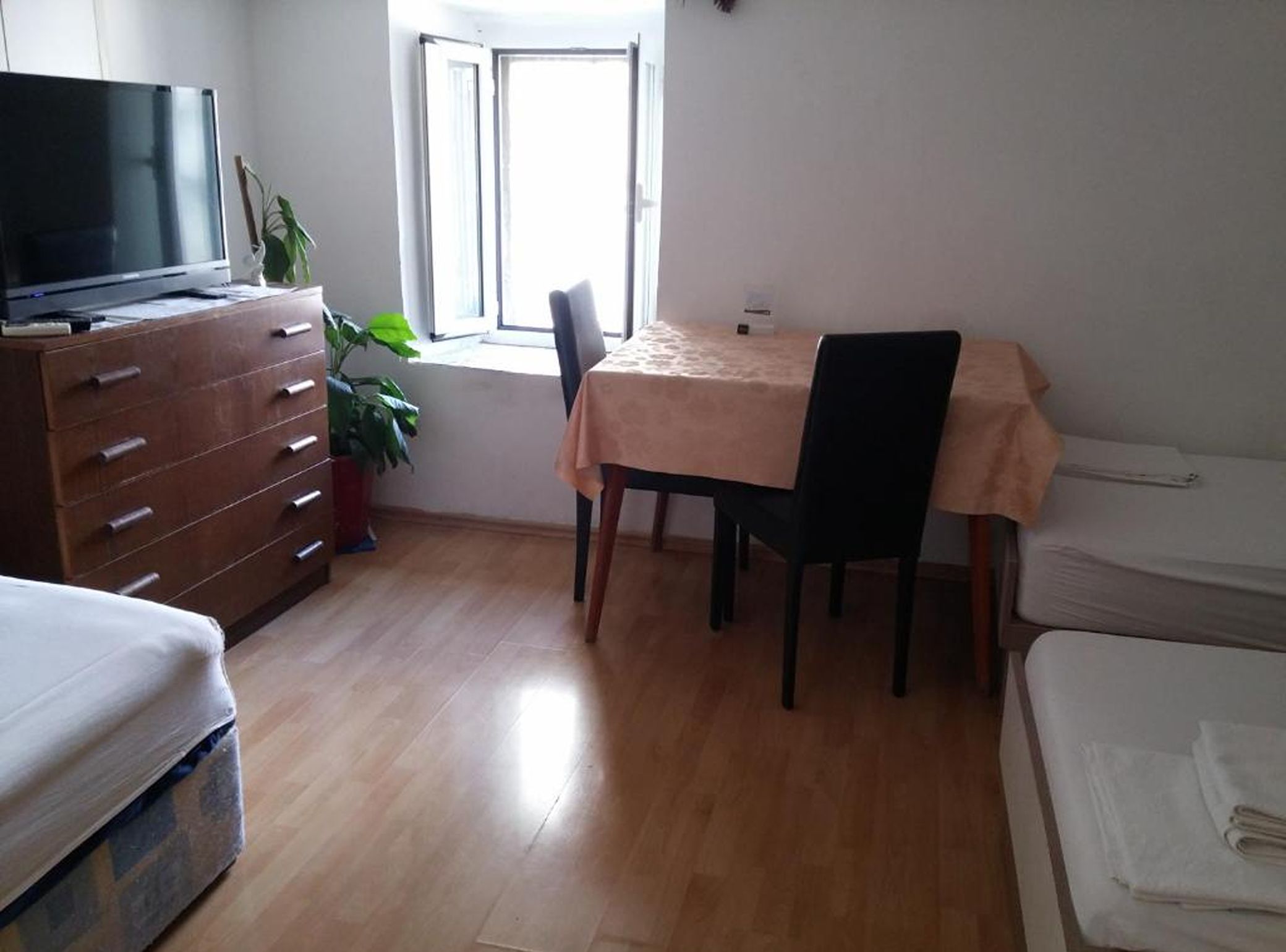 Una habitación con piso de madera, un televisor sobre un mueble, una mesa de comedor con sillas y una ventana con cortinas.