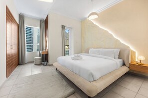 Een moderne slaapkamer met een groot bed, een nachtkastje met lamp, uitzicht op gebouwen door het raam, en een klein krukje.