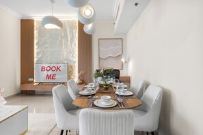 A dining area with a table set for four, a 'BOOK ME' sign, and a large window with curtains. - StayShort - Elegant 2 bedroom that sleeps 4! (JLT)