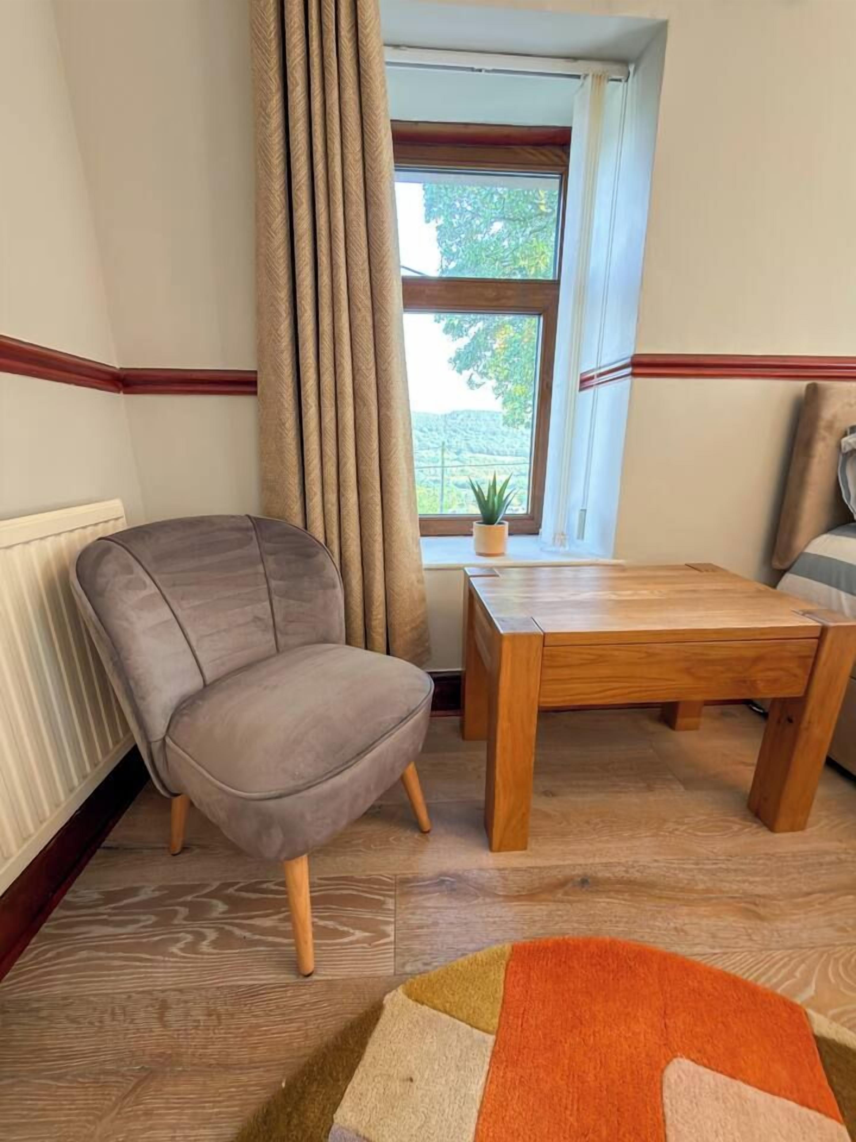 Une pièce comprenant un fauteuil recouvert d’un tissu gris, une table en bois et une fenêtre avec des rideaux.