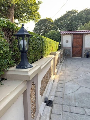 Un chemin pavé mène à un bâtiment doté d’une porte en bois traditionnelle, d’une lanterne noire fixée au mur et d’une haie en arrière-plan.