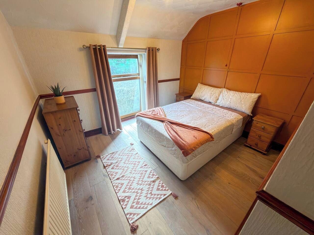 Une chambre à coucher avec un lit, une commode en bois, une fenêtre avec des rideaux et un radiateur.