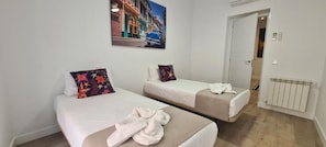Habitación de hotel con dos camas individuales, una pared blanca, un radiador y una puerta que da a otra habitación.