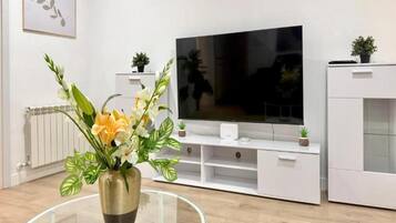 A modern living room with a flat-screen TV, white cabinetry, and a glass coffee table with a floral arrangement.