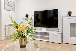 Una sala de estar moderna con un televisor de pantalla plana, muebles blancos y una mesa de centro de vidrio con un arreglo floral.