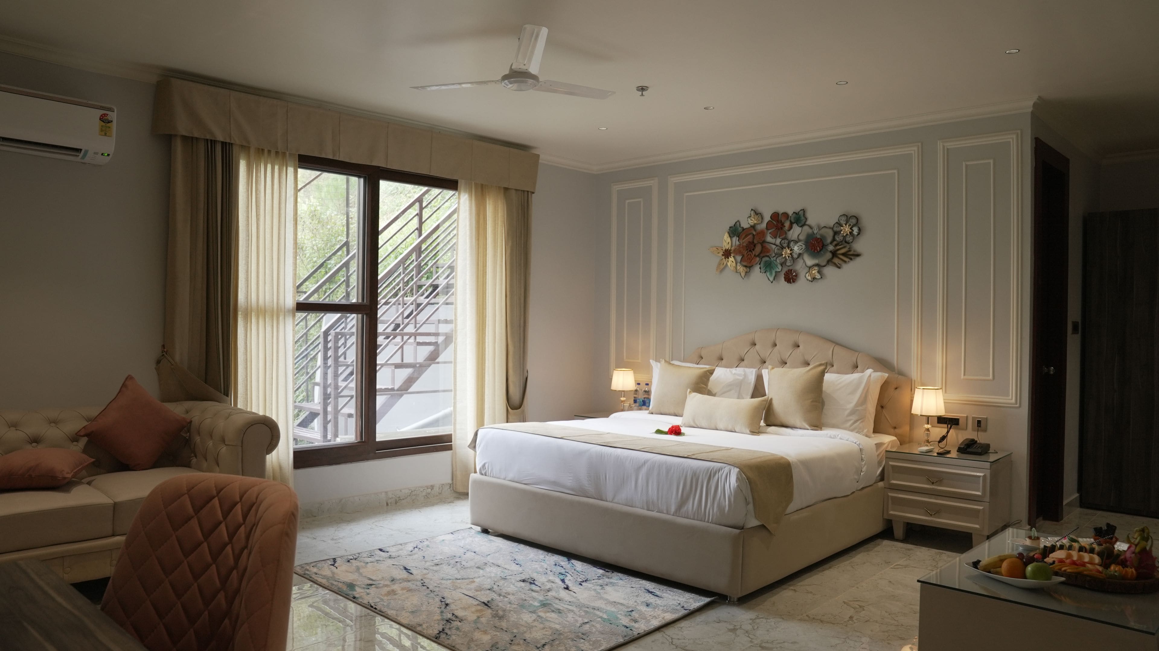 A bedroom with a large bed, a sitting area with a sofa, a dining table with fruit, and a window with a view of a staircase.