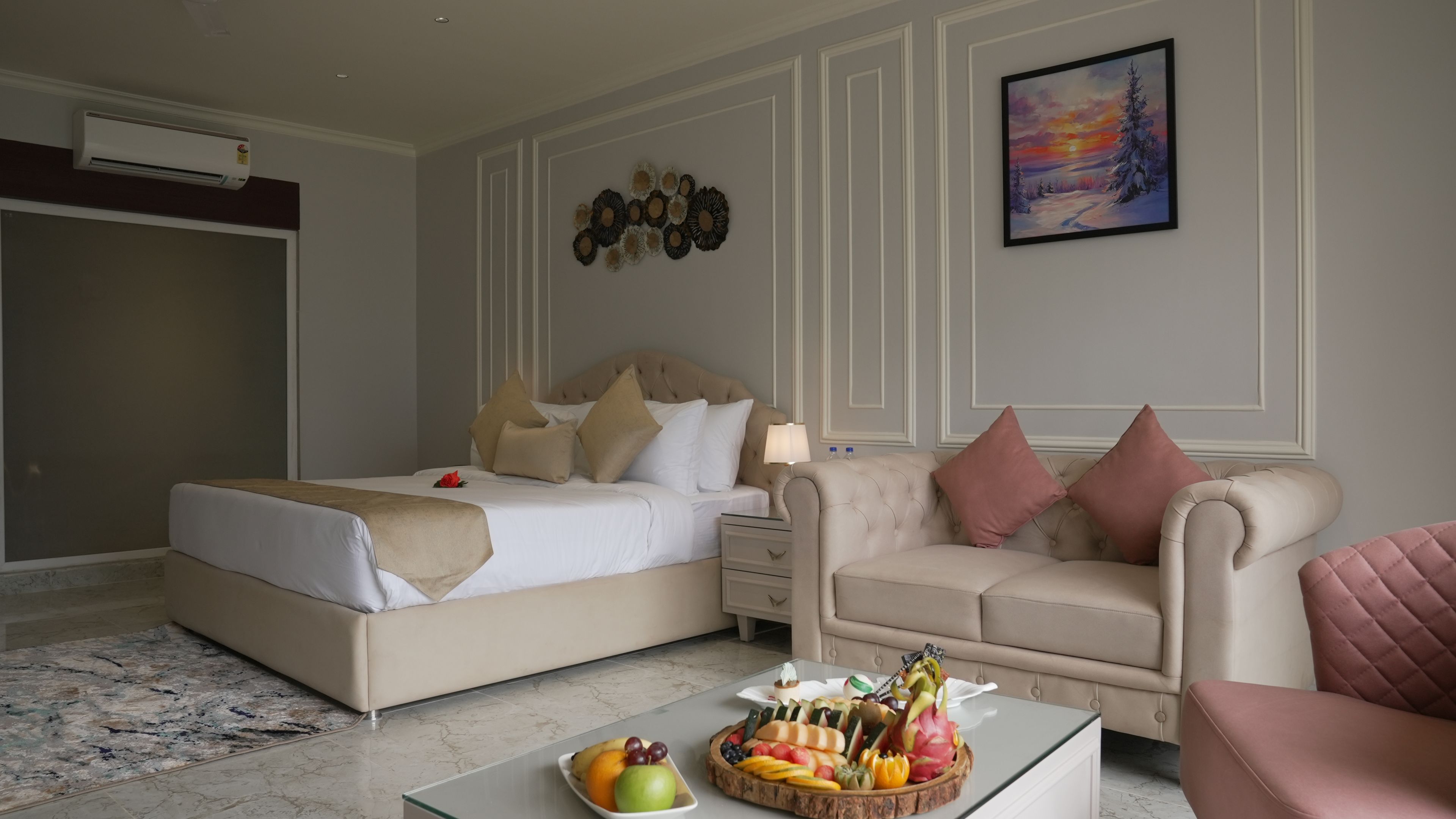 A hotel room with a bed, a sofa, a coffee table with fruit, a painting on the wall, and an air conditioning unit.