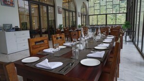 A restaurant with a long dining table set for a meal, wooden chairs, and large windows offering a view of greenery. - Isvara (Kasauli)