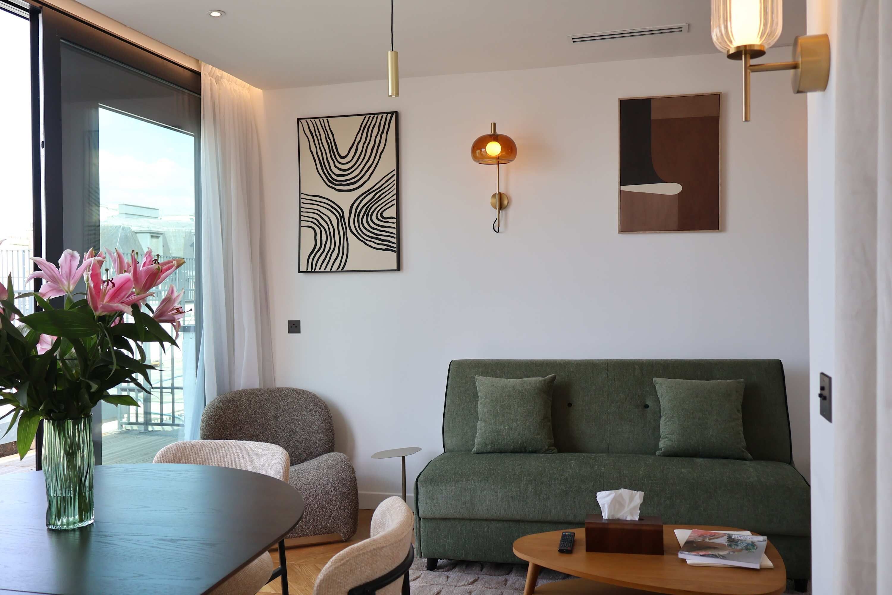 Un salon moderne avec un canapé vert, une table à manger agrémentée d’un vase de fleurs et une grande fenêtre avec des rideaux.