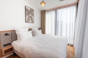 Habitación de hotel con cama, mesita de noche, un cuadro en la pared y una ventana con cortinas translúcidas.