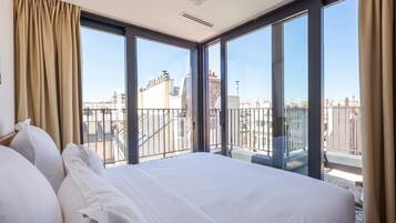Habitación de hotel con una cama grande, vistas de la ciudad a través de ventanales del suelo al techo y un balcón con barandilla.