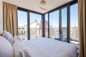 Habitación de hotel con una cama grande, vistas de la ciudad a través de ventanales del suelo al techo y un balcón con barandilla.