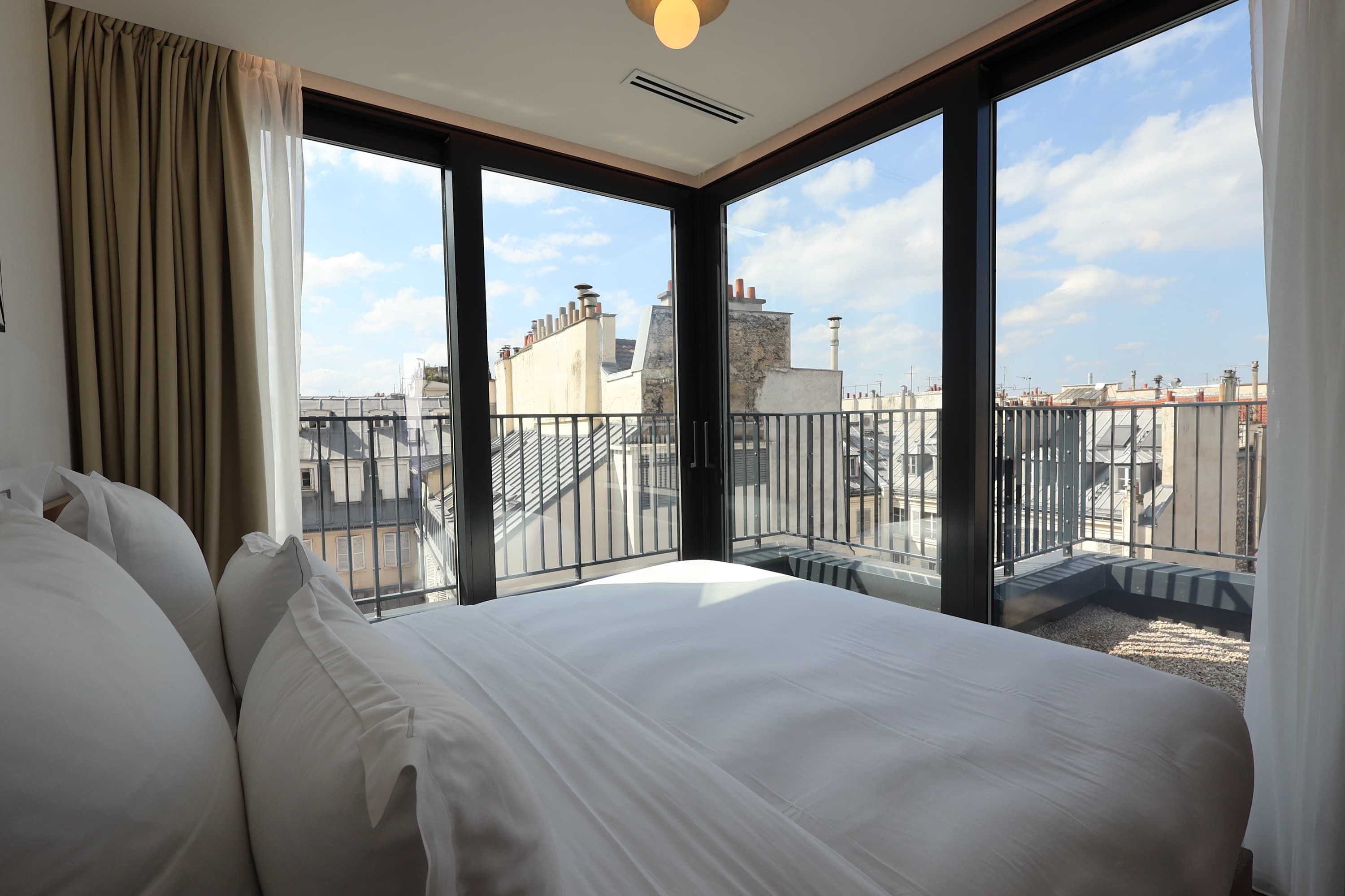 Ein Hotelzimmer mit einem großen Bett, Blick auf die Stadt durch raumhohe Fenster und einem Balkon mit Metallgeländer.
