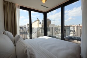 Ein Hotelzimmer mit einem großen Bett, Blick auf die Stadt durch raumhohe Fenster und einem Balkon mit Metallgeländer.
