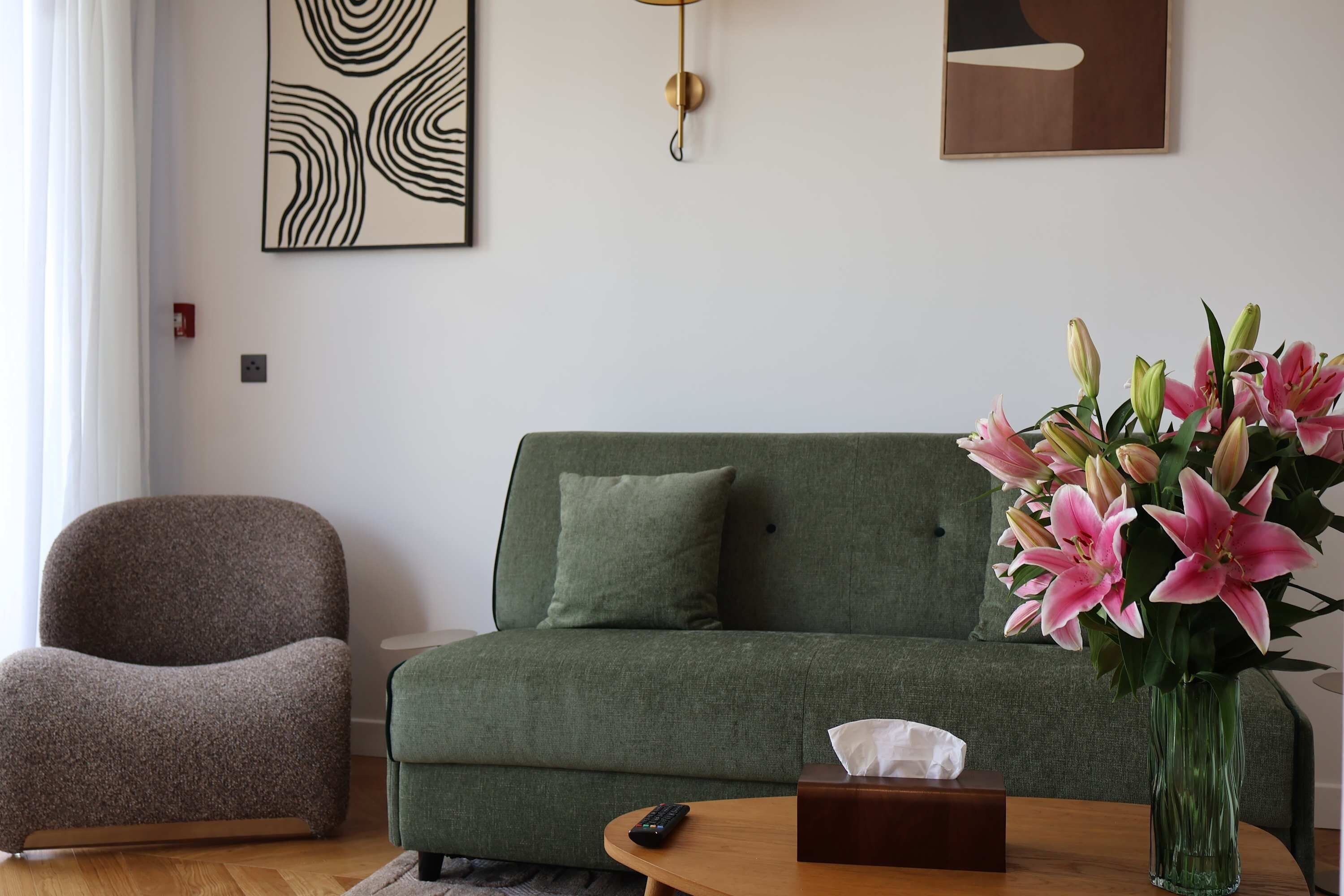 Una sala de estar moderna con un sofá verde, un sillón marrón, un jarrón de lirios rosados y un control remoto sobre una mesita de madera.