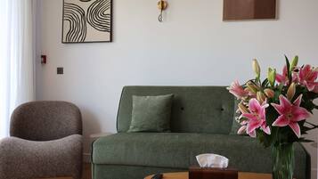 Una sala de estar moderna con un sofá verde, un sillón marrón, un jarrón de lirios rosados y un control remoto sobre una mesita de madera.