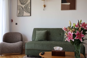 Ein modernes Wohnzimmer mit einem grünen Sofa, einem braunen Sessel, einer Vase mit rosa Lilien und einer Fernbedienung auf einem Holztisch.