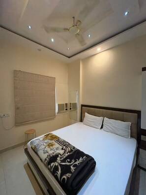 A hotel room with a bed, a ceiling fan, recessed lighting, a mirror, and a window with blinds. - Saraswati Grand (Varanasi)