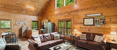 A cozy cabin interior with a wooden ceiling, a dining area, a living room with leather sofas, and a kitchen area.