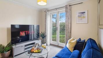 En stue med et fladskærms-tv, en blå sofa, et sofabord med frugt og et glas, samt en balkon med udsigt.