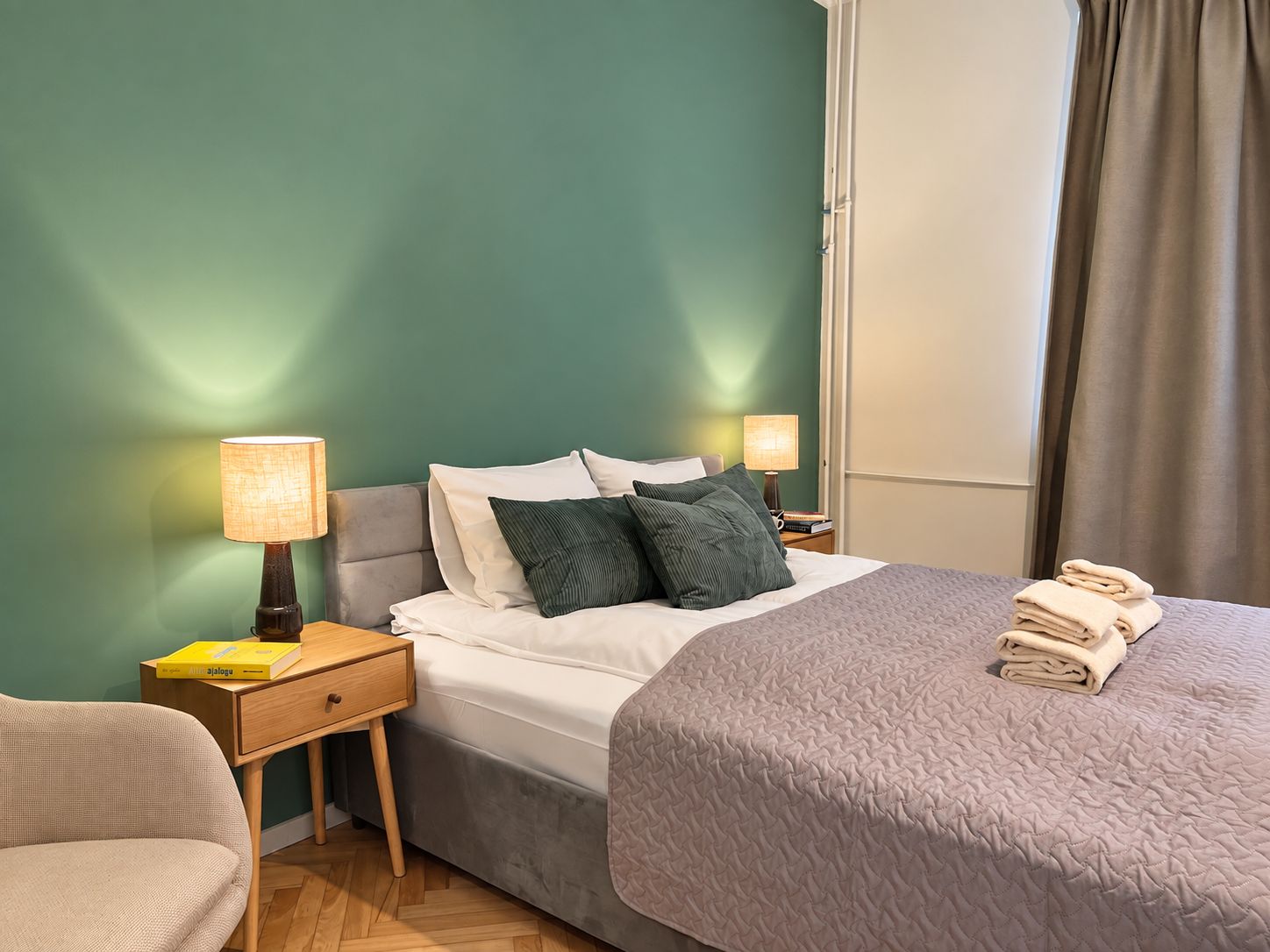 Ein Hotelzimmer mit einem Bett, Nachttischen, Lampen, einem Sessel und einem Fenster mit Vorhängen.