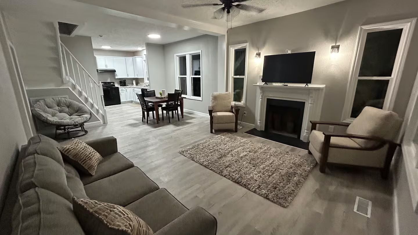 A living room with a sofa, chairs, a fireplace, a television, and a kitchen in the background.