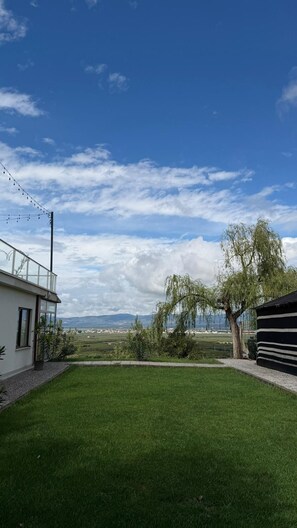 A grassy yard with a view of a lake and mountains, a tree, and a building with a balcony.