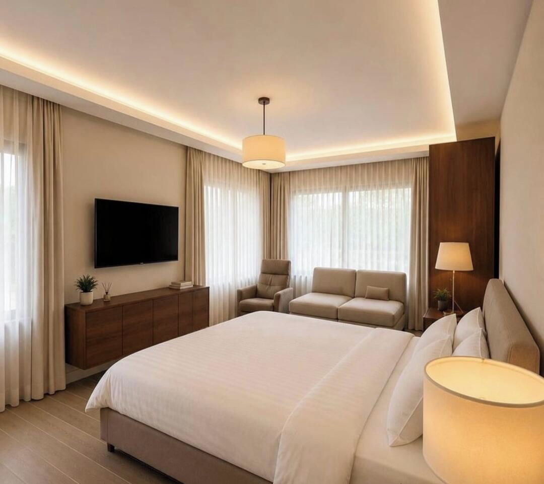 Habitación de hotel moderna con una cama grande, un televisor de pantalla plana, un aparador de madera, una zona de estar con un sofá y sillones, y una mesita de noche con una lámpara.
