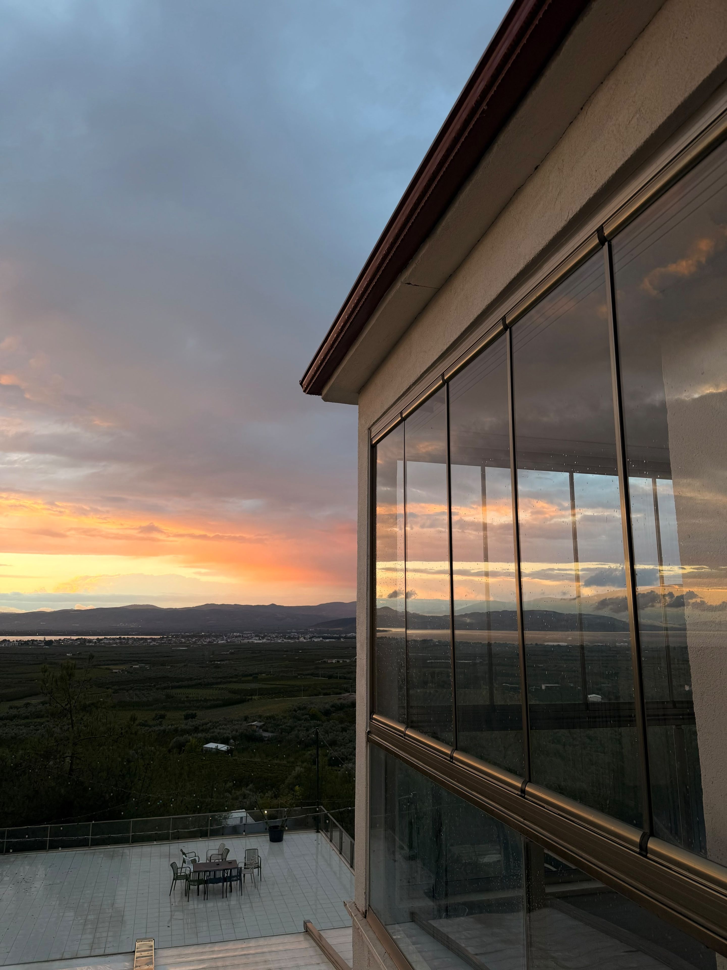 Ein Balkon mit Blick auf einen Sonnenuntergang über einer Landschaft, darunter Hügel in der Ferne und ein Gewässer.