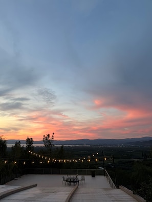 Una terraza al aire libre con una mesa y sillas, con vistas a un paisaje con montañas lejanas y un cielo al atardecer.