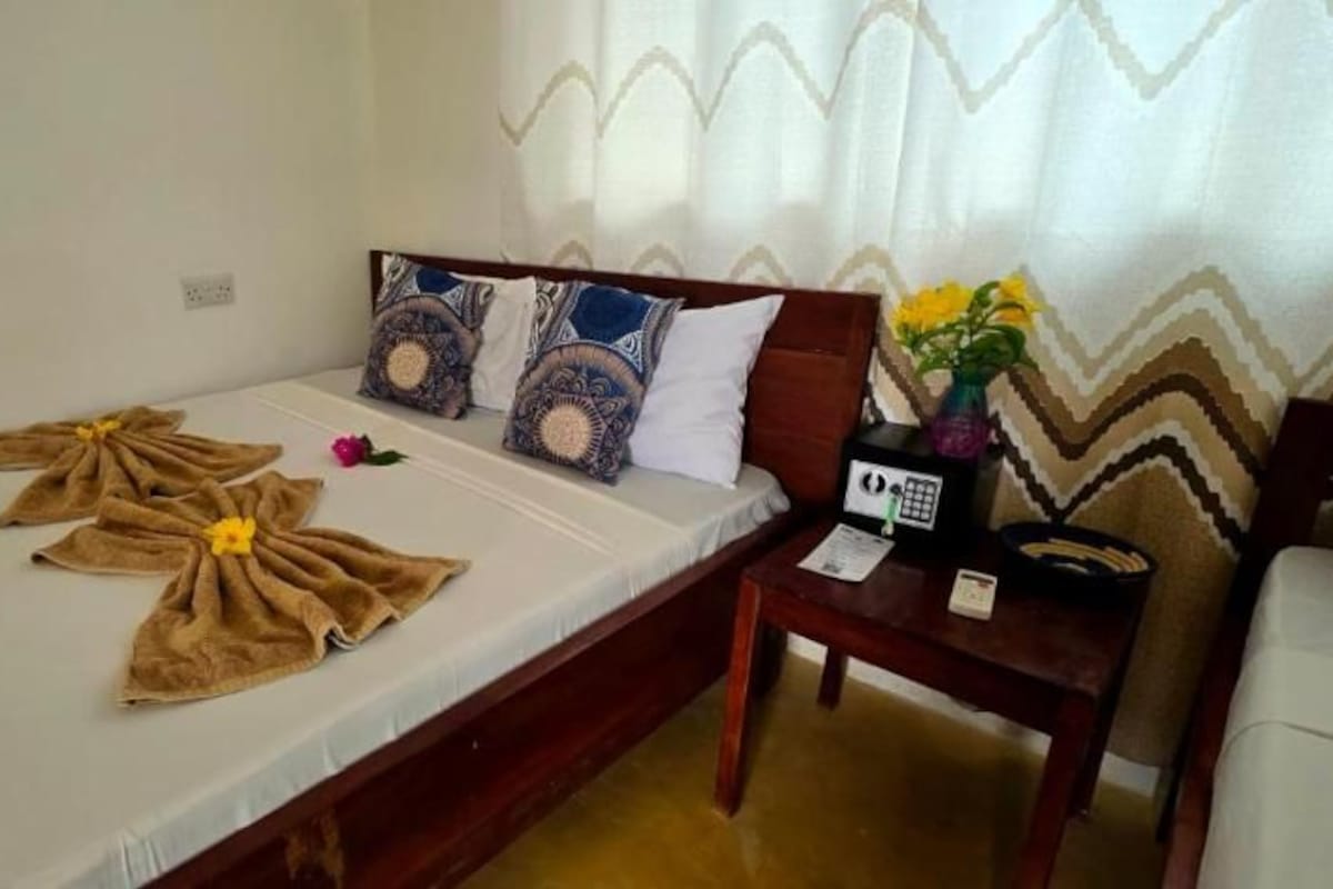 Una cama individual con colchón blanco, cabecera de madera y almohadas estampadas. Una mesita de noche con un teléfono y un florero está al lado de la cama.