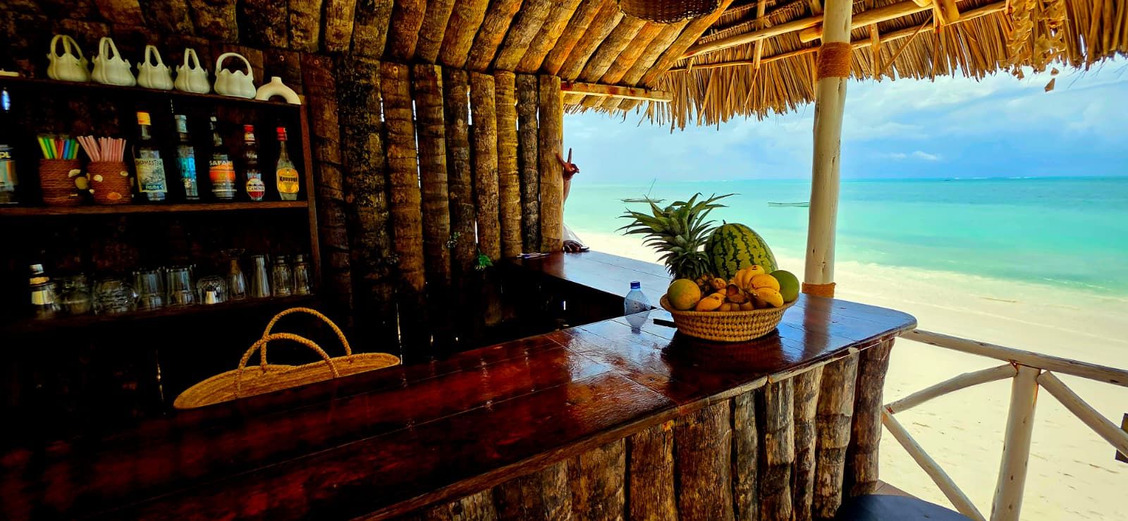 A beachside bar with a wooden counter, a thatched roof, and a view of the ocean.
