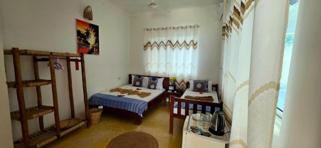 A room with two beds, a wooden shelf, a small table, and a window with curtains.