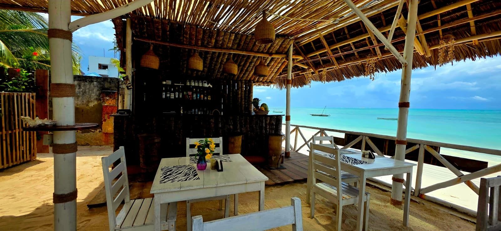 Un bar frente al mar con techo de paja, mesas y sillas blancas, y vistas al océano.