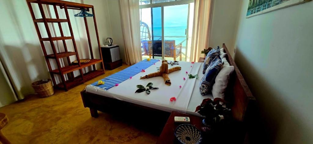 Une chambre avec un lit, une étagère en bois, une petite table avec une lampe et une vue sur la mer, visible à travers une porte vitrée.