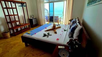 Une chambre avec un lit, une étagère en bois, une petite table avec une lampe et une vue sur la mer, visible à travers une porte vitrée.