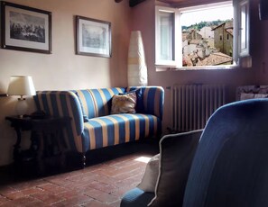 Ein Wohnzimmer mit einem gestreiften Sofa, einer Lampe, einem Heizkörper und einem Fenster mit Blick auf Gebäude.