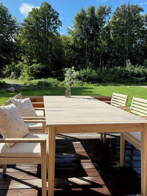 Eine Holzterrasse im Freien mit Tisch und Bänken, umgeben von Grünpflanzen und Bäumen.