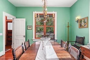 Uma sala de jantar com uma mesa de madeira comprida, cadeiras, um candelabro e uma janela grande com vista para vegetação.