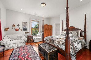Ein Bett mit Baldachin, mit einem gemusterten Bettbezug, eine geschnitzte Holzbank, ein weißes Sofa und ein Teppich in Rot und Schwarz in einem Schlafzimmer mit Holzboden und einem großen Fenster.