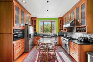 A modern kitchen with wooden cabinets, stainless steel appliances, a central island with stools, and a large window with a view of trees. - Napa Extended Stay | 7-Acre Vineyard Farmhouse (Napa)